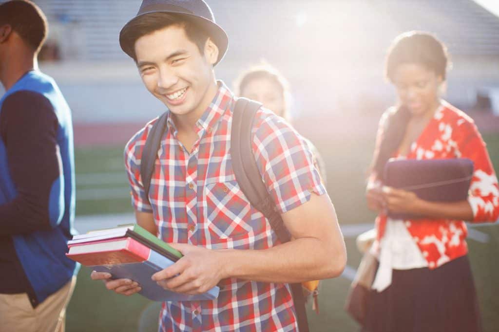 A yound college student in a plaid shirt carries his books and smiles laughingly with his friends