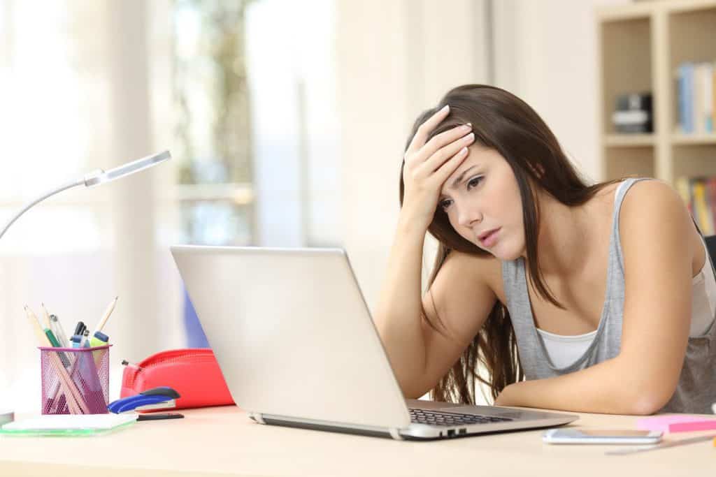 A high school girl leaning over her laptop, her palm pressed to her forehead and her face painted with exasperation