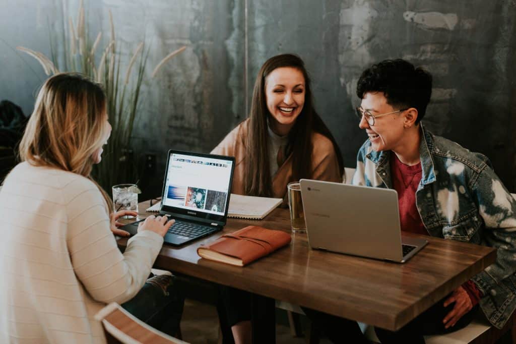 Three college-aged friends sitting around a table working at their laptops and laughing