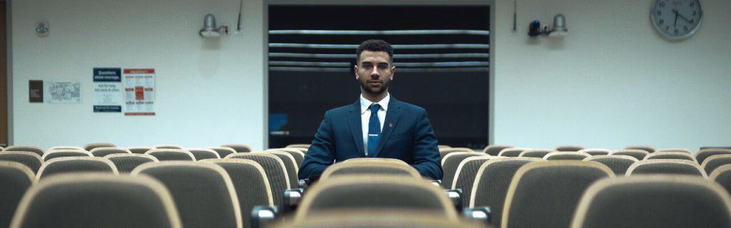 A man in a suit and tie sits at the center of a dim lecture hall, surrounded by empty seats