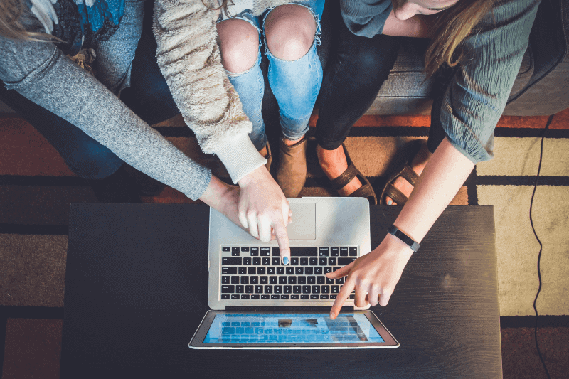 3 students huddle around a laptop at a comfortable cafe and point at elements of a college finder quiz